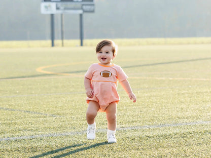 Toddler boy in an orange striped football shirt with a football graphic on a sunlit grassy field.