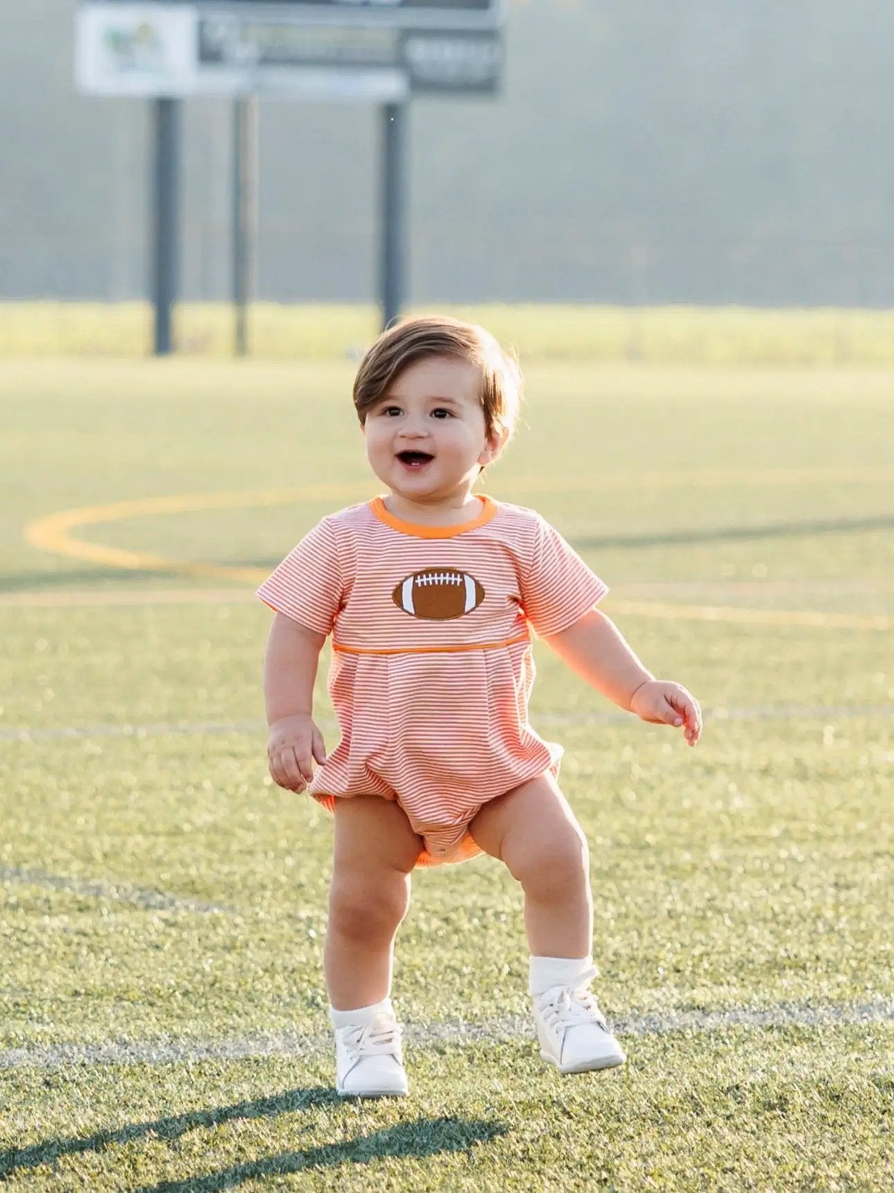 Smiling toddler boy in orange striped football bubble romper on a grassy field.