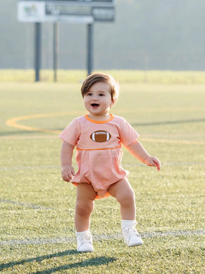 Smiling toddler boy in orange striped football bubble romper on a grassy field.