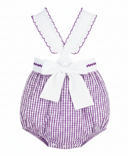 A baby romper with purple and white gingham patterned bloomers, white shoulder straps with purple zigzag trim, and a large white bow at the waist.